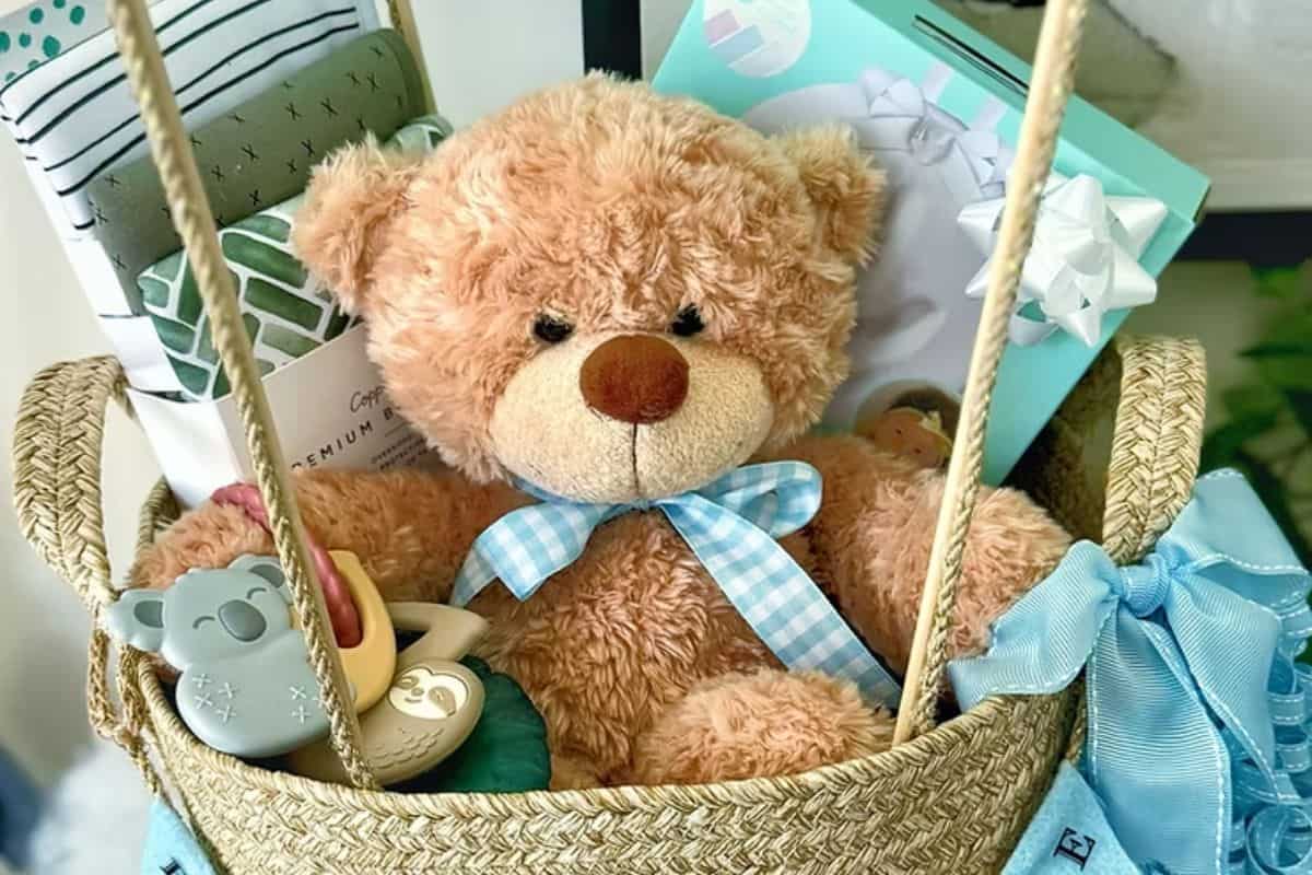 Woven baby shower gift basket filled with a teddy bear, baby blankets, toys, and gift-wrapped items, tied with a blue ribbon.