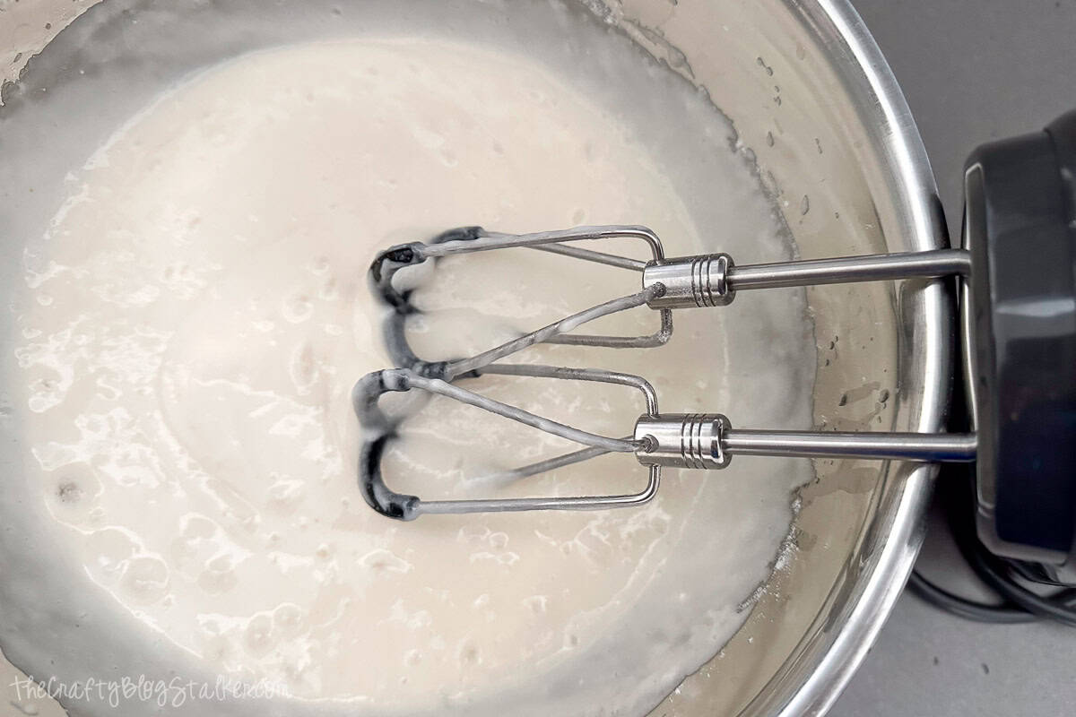 Electric hand mixer with beaters mixing smooth white cake batter in a stainless steel bowl.