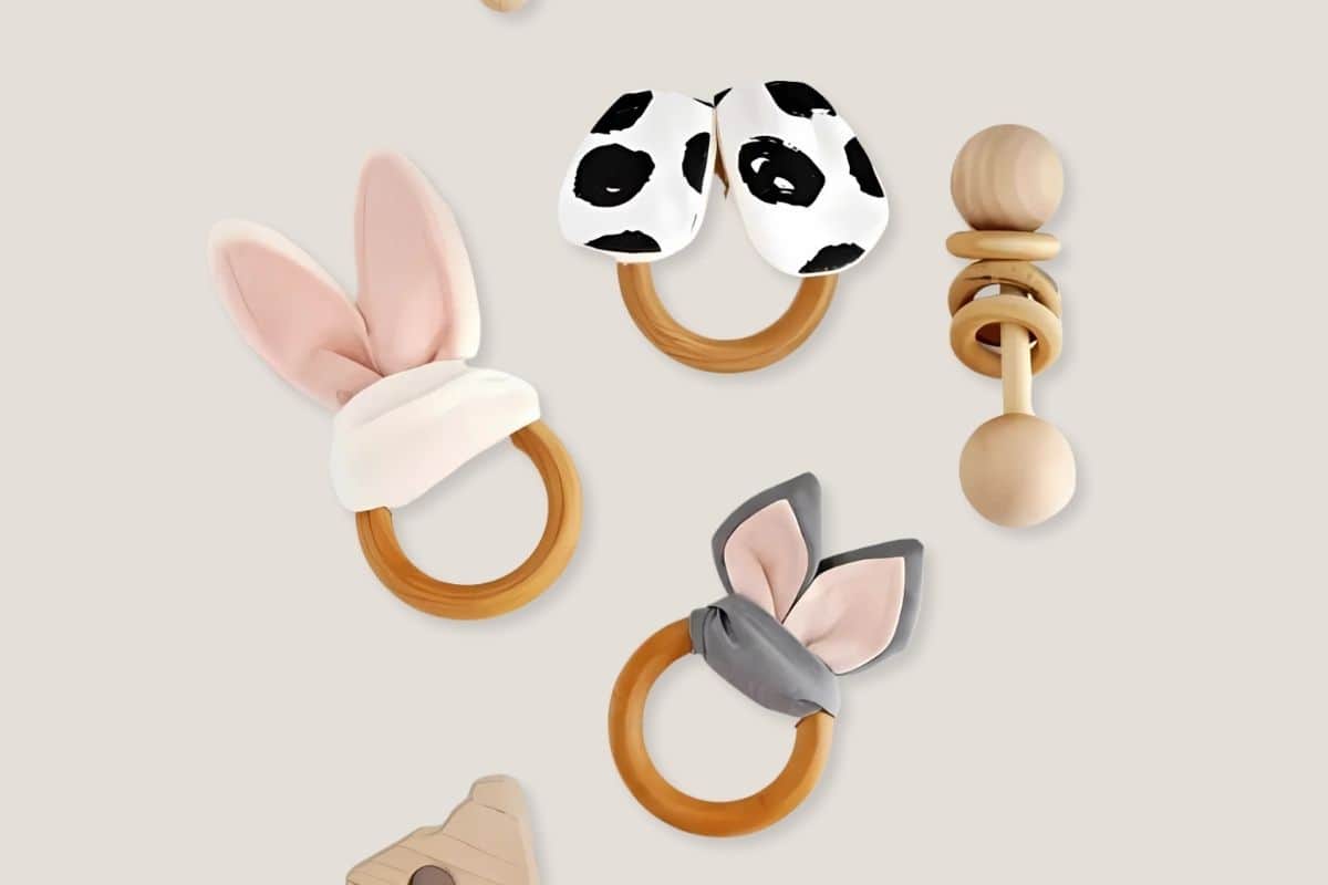 Assorted wooden baby teethers with soft fabric ears in bunny and cow prints, plus a natural wooden rattle, all on a neutral background.