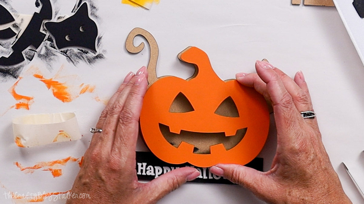 Hands aligning a painted orange jack-o’-lantern cutout onto a wooden Halloween base with the words “Happy Halloween” visible at the bottom.