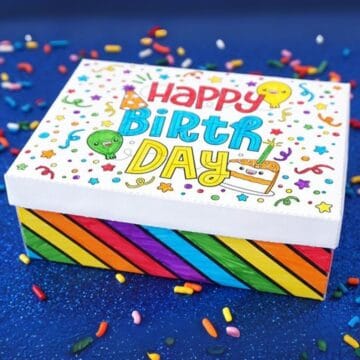 Colorful DIY birthday gift box with "Happy Birthday" lid design, rainbow-striped sides, and confetti sprinkles on a blue background.