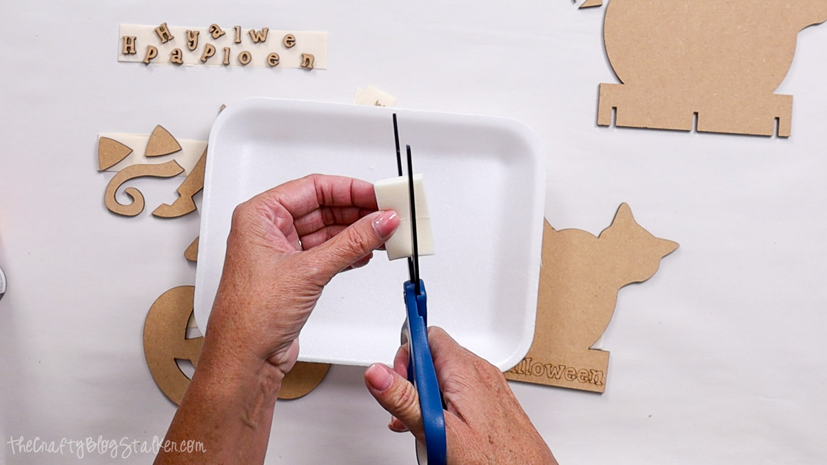 Hands cutting a makeup sponge with scissors over a white tray, preparing supplies for painting small laser-cut Halloween craft pieces.