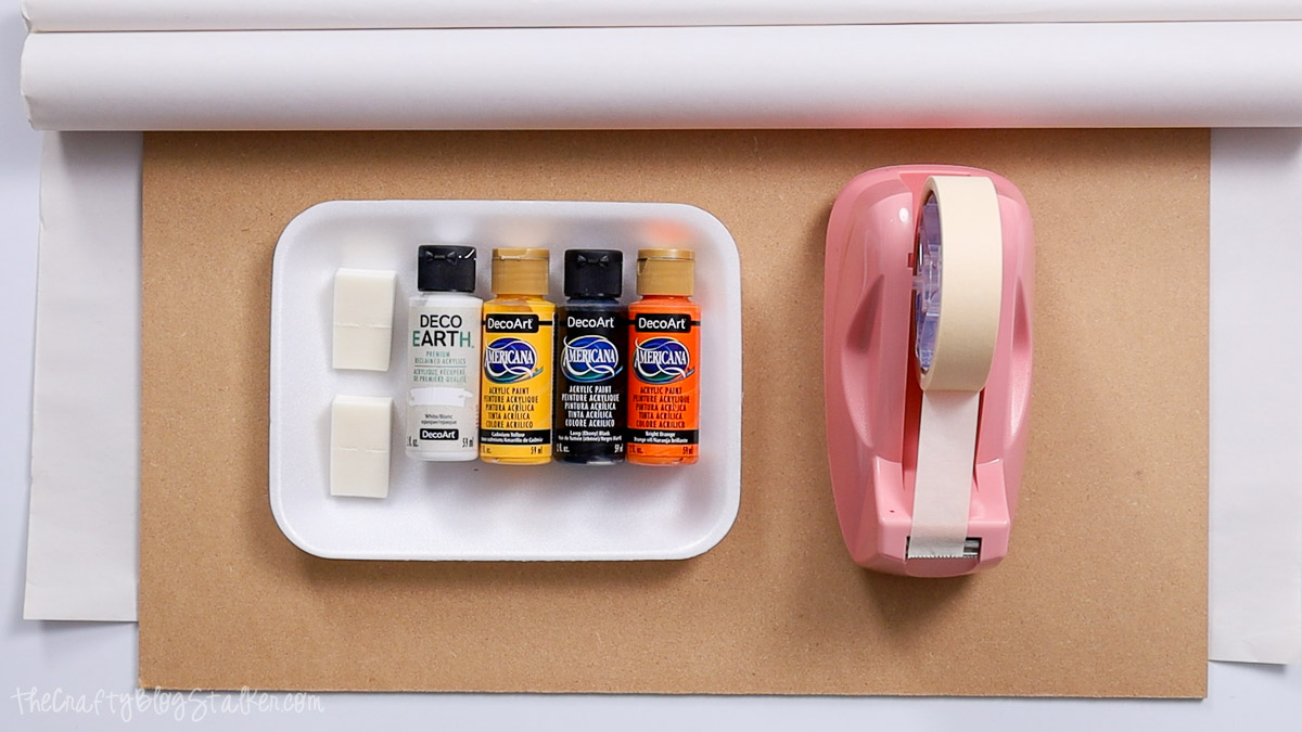 Craft supplies laid out including acrylic paints, foam brushes, masking tape dispenser, white paper, and a brown board for a DIY Halloween project.