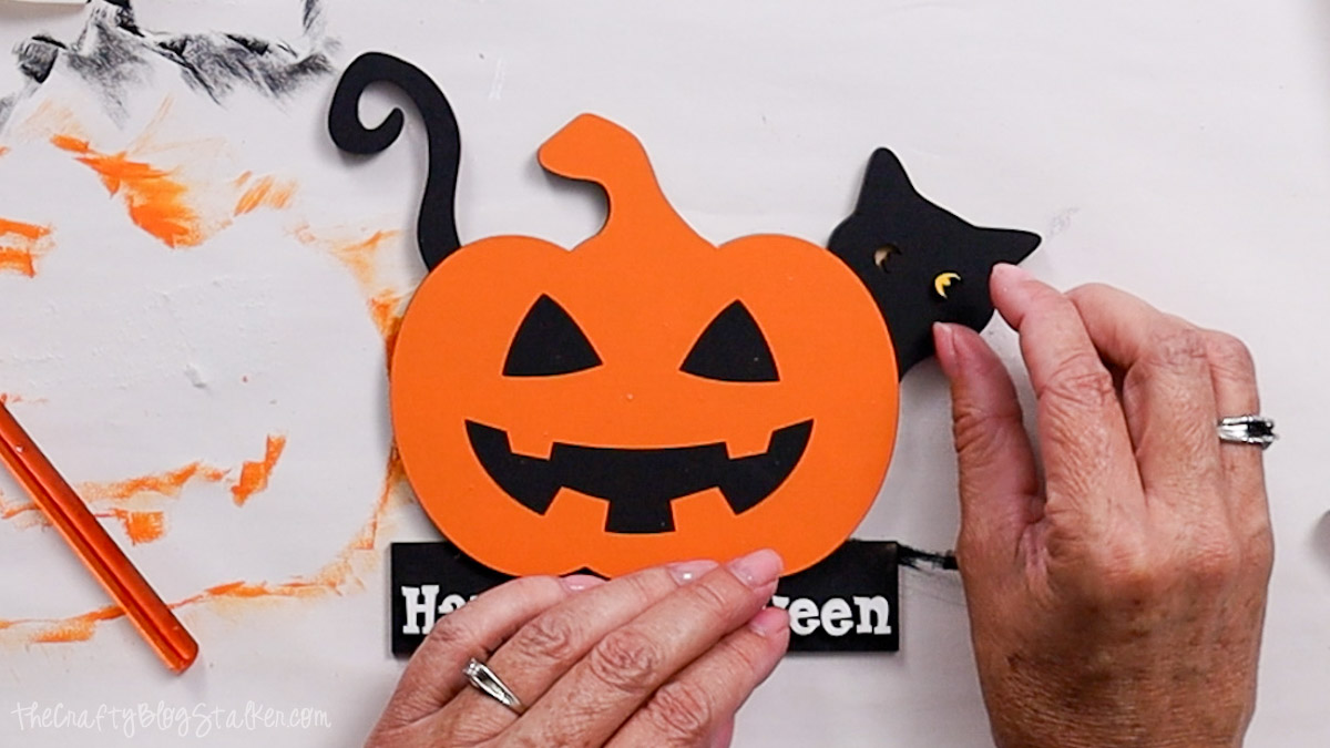 Hands assembling a Halloween craft by gluing black cat pieces with yellow eyes behind an orange jack-o’-lantern cutout on a “Happy Halloween” base.