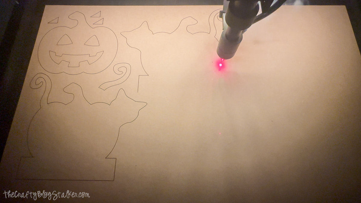 Laser cutting machine cutting Halloween shapes of a pumpkin, cat, and text onto wood, preparing pieces for a DIY Halloween mantel decor project.
