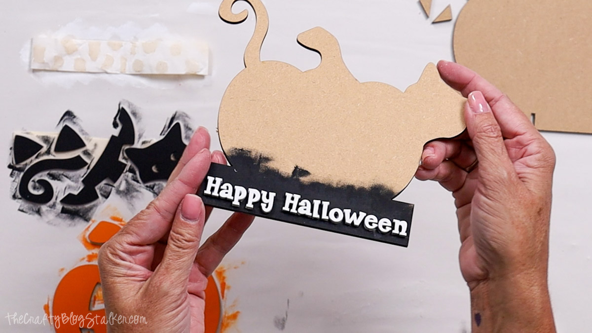Hands holding a laser-cut wooden Halloween decoration with a cat shape and painted black base featuring the words "Happy Halloween" in white letters.
