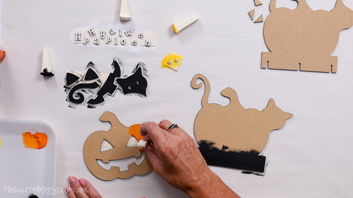 Hand painting a laser-cut wooden pumpkin orange with a sponge, while black-painted Halloween cat and lettering pieces dry on the table.