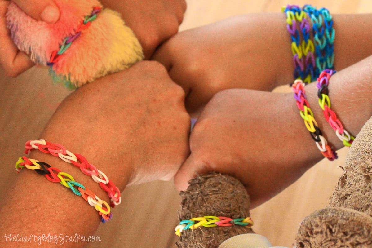 A group of hands and arms showing off colorful loom band bracelets in different patterns, some worn by kids and some on stuffed animals.
