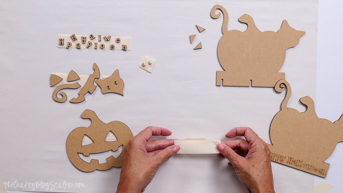 Hands organizing and taping laser-cut MDF Halloween pieces, including pumpkin, cat, letters, and small details, for a DIY Halloween craft project.