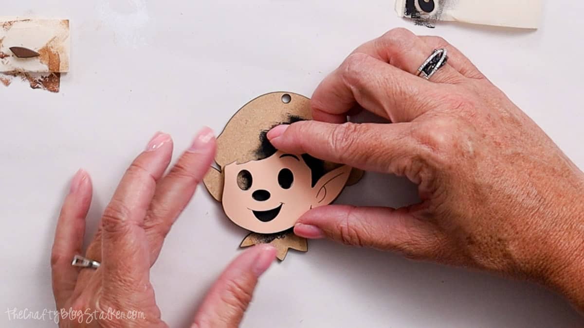 Hands gluing a painted elf face cutout onto a wooden base ornament, showing the elf's smiling expression with ears and facial features.