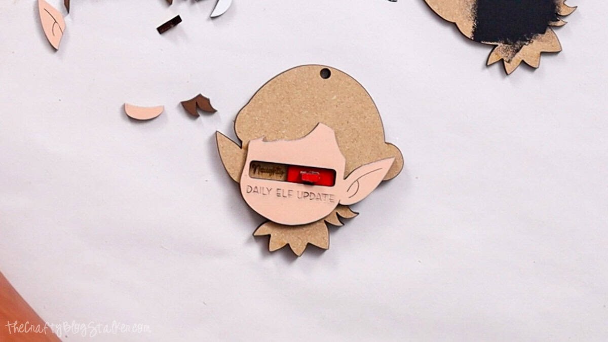 Partially assembled laser cut elf ornament with face piece glued over a red slider showing "Naughty/Nice" and text "Daily Elf Update" at the bottom.