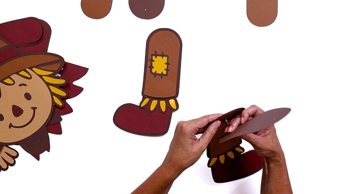Hands attaching the upper part of a paper scarecrow leg with a brad fastener, next to a completed leg with a red boot, yellow fringe, and a patch detail.