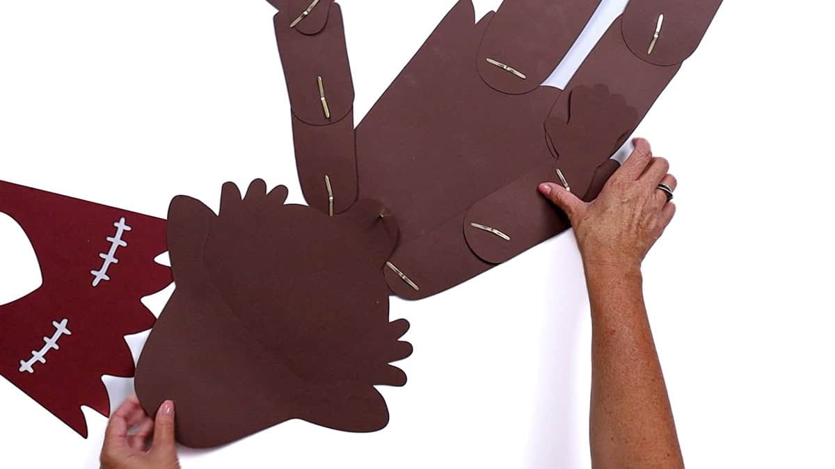 Hands attaching the scarecrow head to the paper body using brad fasteners, with the arms and legs already secured and the red shirt piece set aside.