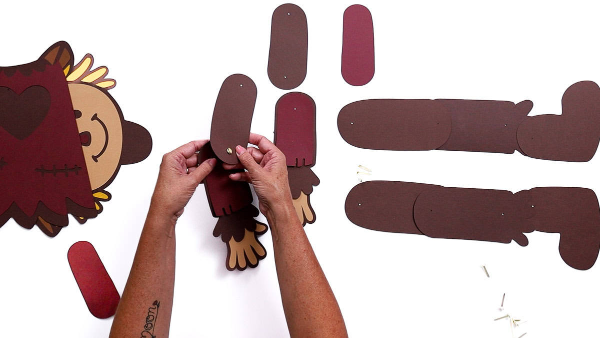 Hands attaching the upper arm piece to a paper scarecrow using a brad fastener, with other cutout body parts laid out for assembly.