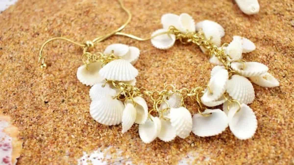 DIY seashell bracelet with white shells attached to a gold chain, displayed on sandy beach background for a coastal-inspired accessory.
