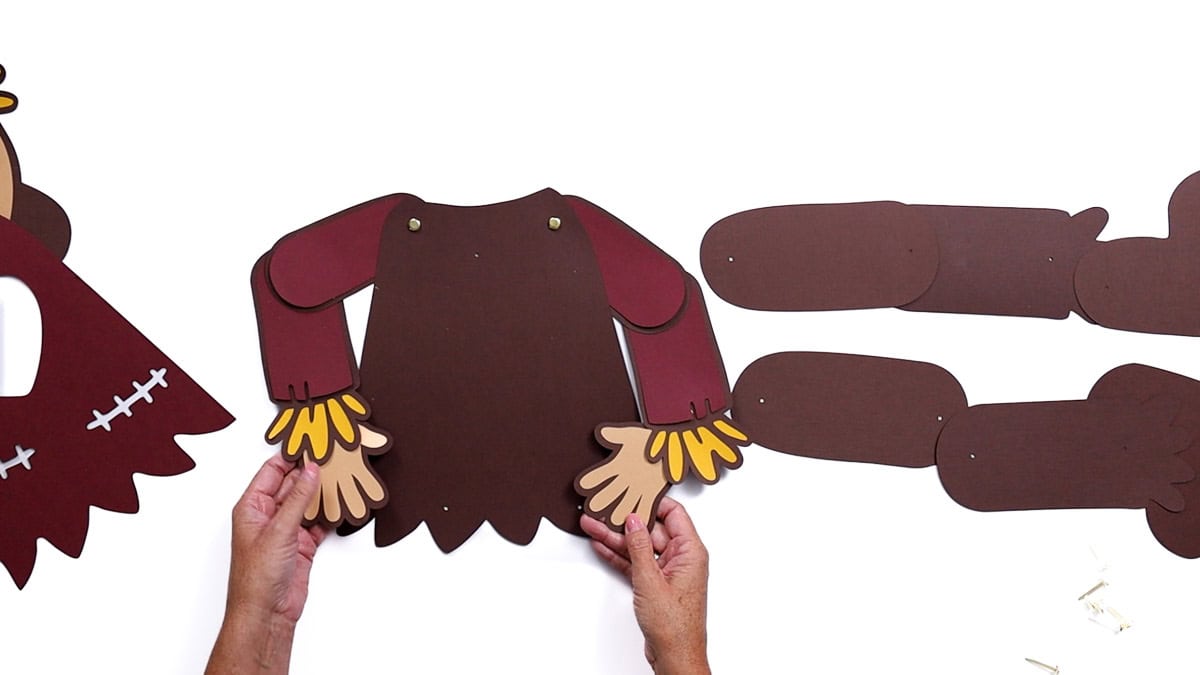 Hands holding a completed paper scarecrow body with red sleeves, tan hands, and yellow fringe, assembled with brad fasteners.
