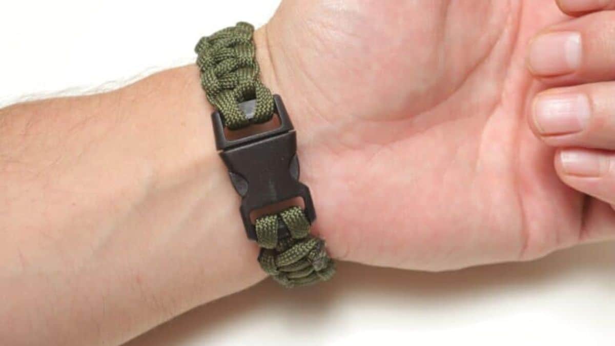 A green paracord survival bracelet with a black plastic buckle is shown worn on a wrist, highlighting its braided design and sturdy clasp.