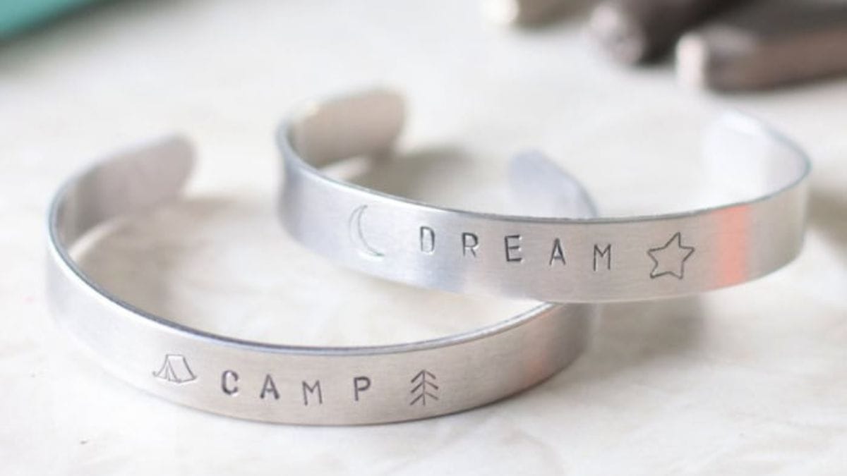 Two silver metal stamped cuff bracelets, one engraved with "CAMP" and outdoors icons, the other with "DREAM" and celestial symbols.