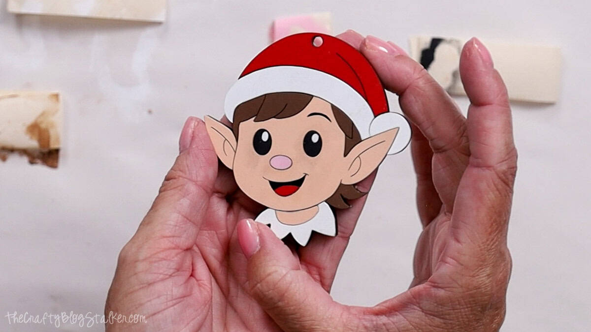 Hands holding a finished elf face ornament with a red Santa hat, big black eyes, pink nose, and white collar, ready to hang as Christmas decor.