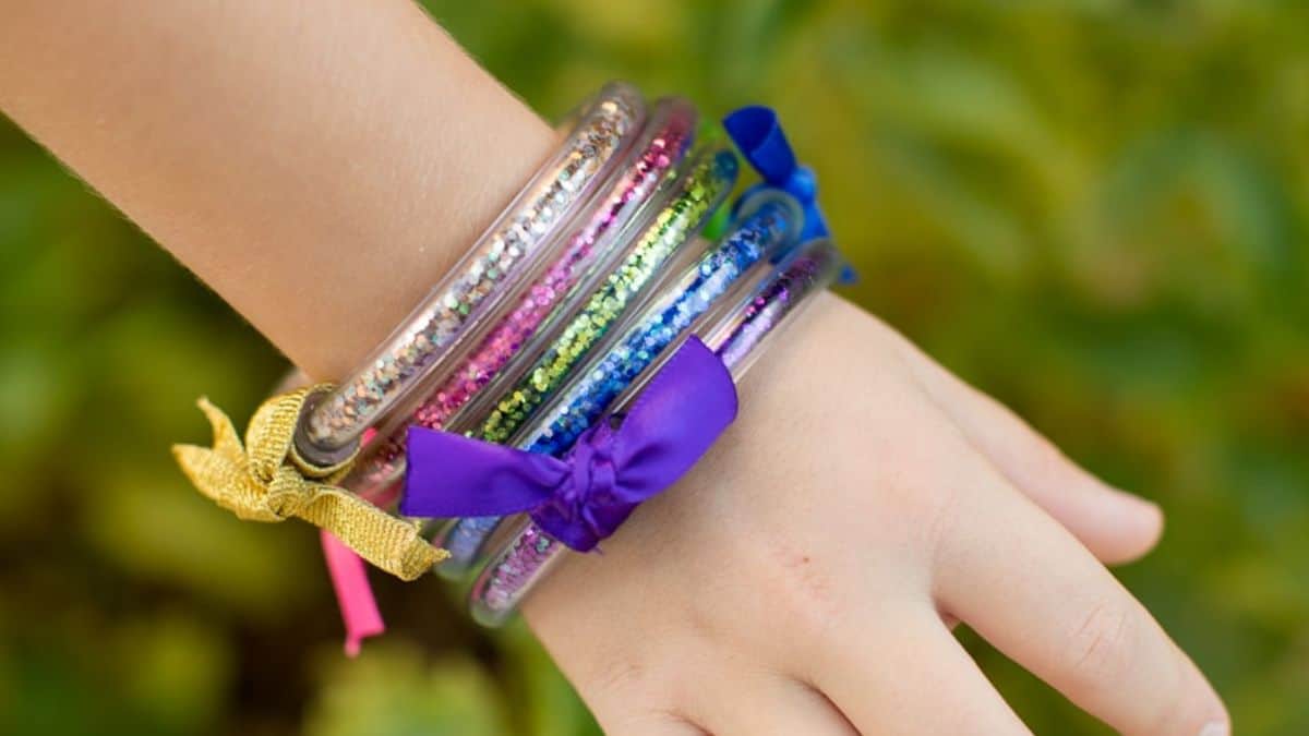 Child wearing colorful DIY glitter tube bracelets tied with ribbons in purple, blue, pink, and gold, sparkling in the sunlight.