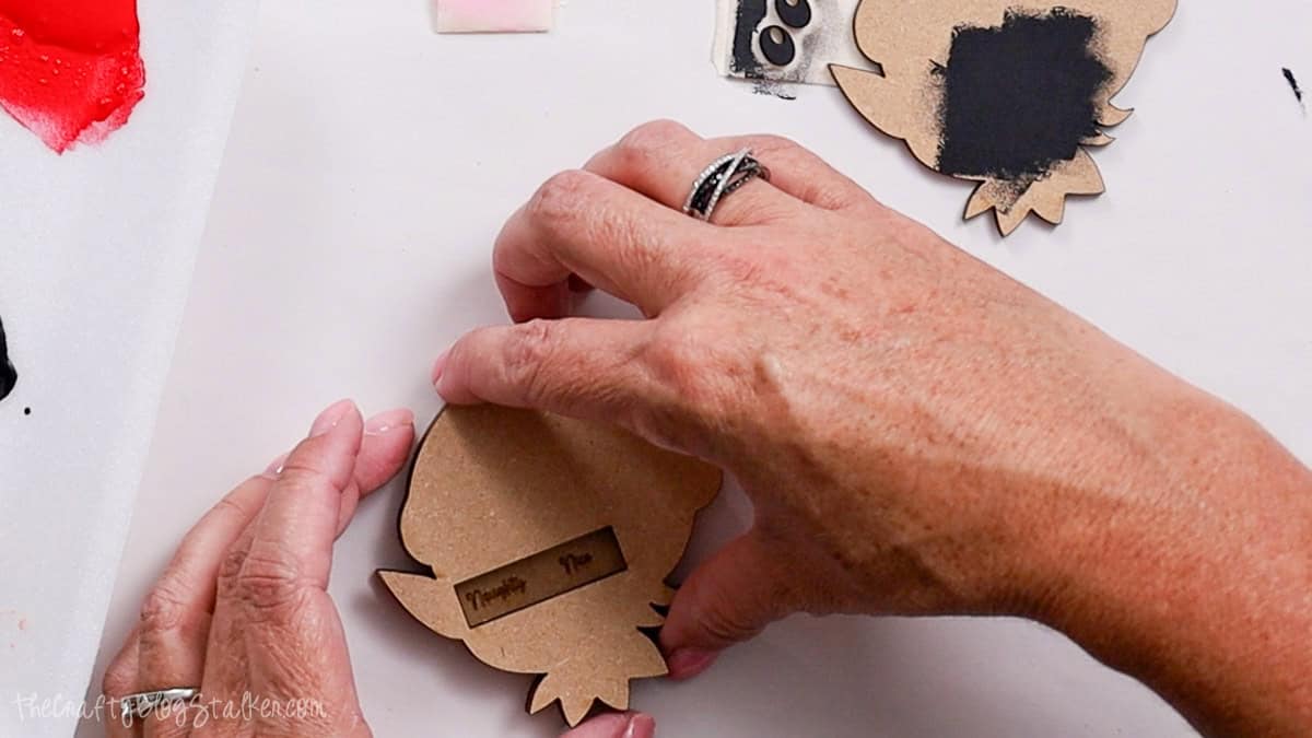 Hands gluing together base layers of a laser cut wood elf ornament, with "Naughty" and "Nice" text visible on the back piece.