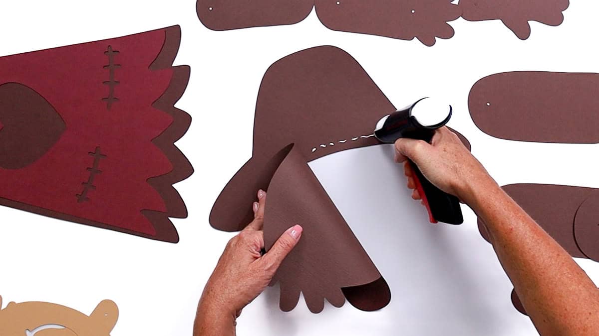 Hands gluing together brown paper hat pieces with a glue gun as part of assembling a DIY scarecrow craft project.