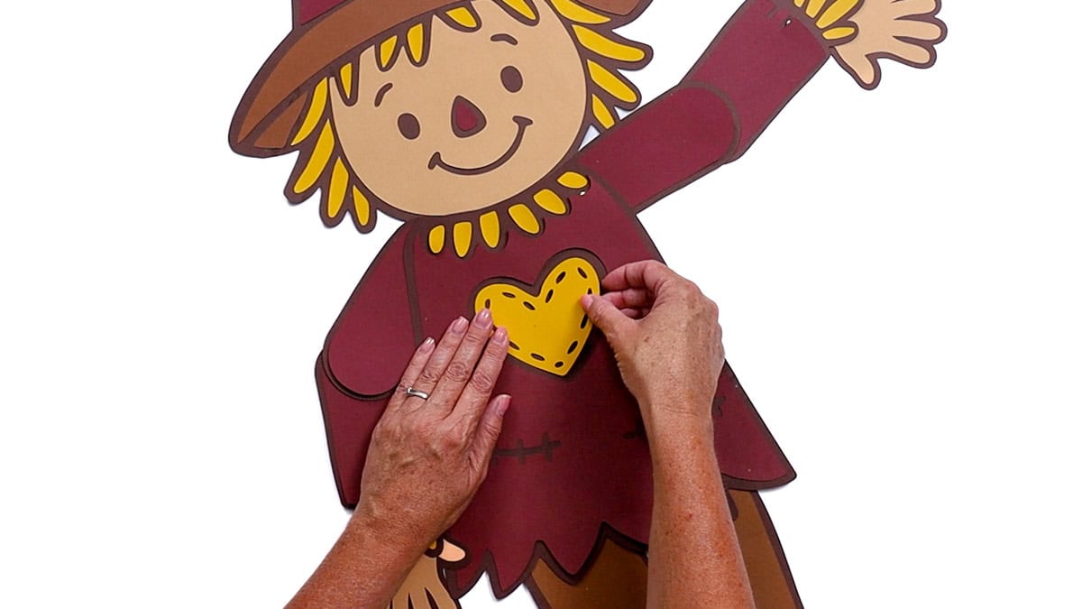 Hands gluing a yellow heart onto the chest of a completed paper scarecrow with movable arms, legs, and a smiling face wearing a brown hat.