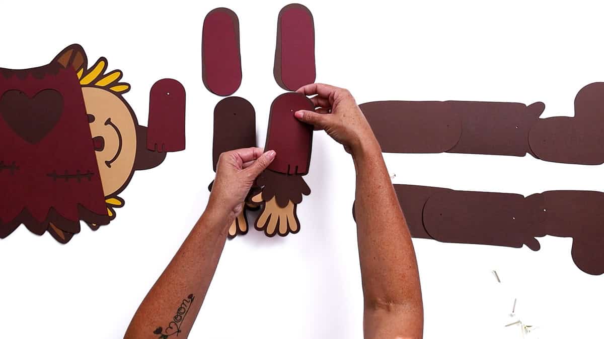 Hands attaching a red paper sleeve to the arm of a paper scarecrow craft, with other cutout body parts laid out for assembly.