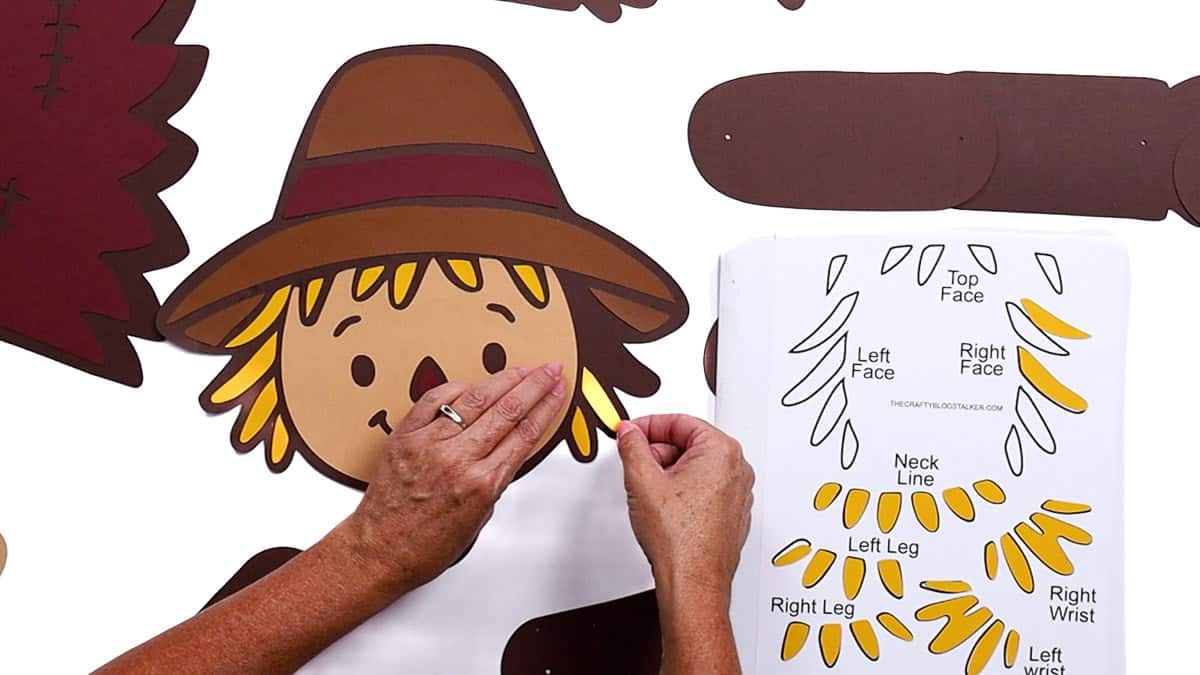 Hands attaching yellow paper hair fringe pieces to a scarecrow face cutout, using a placement guide for accurate assembly in a DIY craft project.