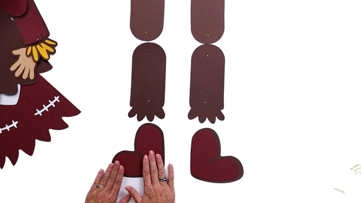 Hands gluing red boot cutouts onto brown paper pieces while assembling the legs of a paper scarecrow craft.