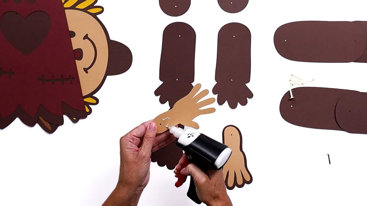 Hands applying glue to attach tan paper hand cutouts for a scarecrow craft, with other arm and leg pieces spread out on the table for assembly.