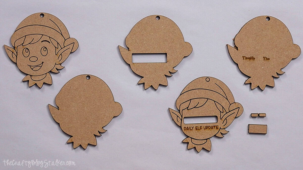 Laser cut wood pieces shaped like elf faces, including engraved details and cutouts, ready to assemble into Christmas ornaments with a "Daily Elf Update" design.