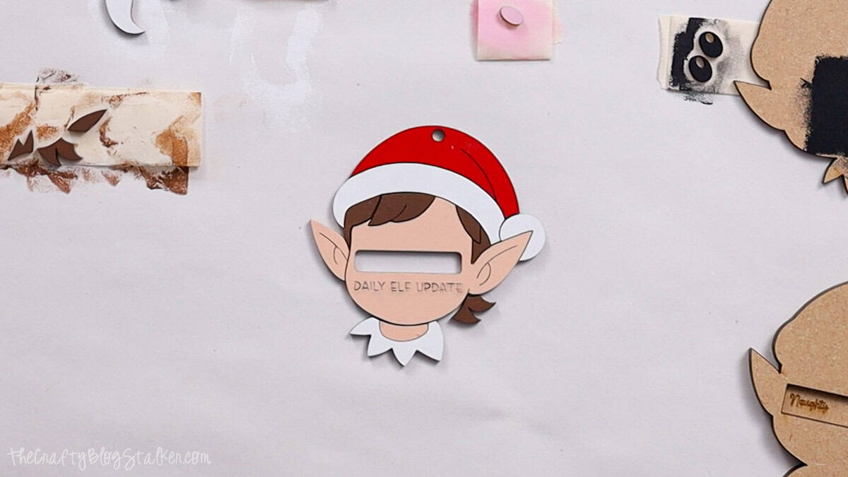 Partially assembled laser cut elf ornament with a red Santa hat, white collar, ears, and hair, showing a cut-out slot labeled "Daily Elf Update."