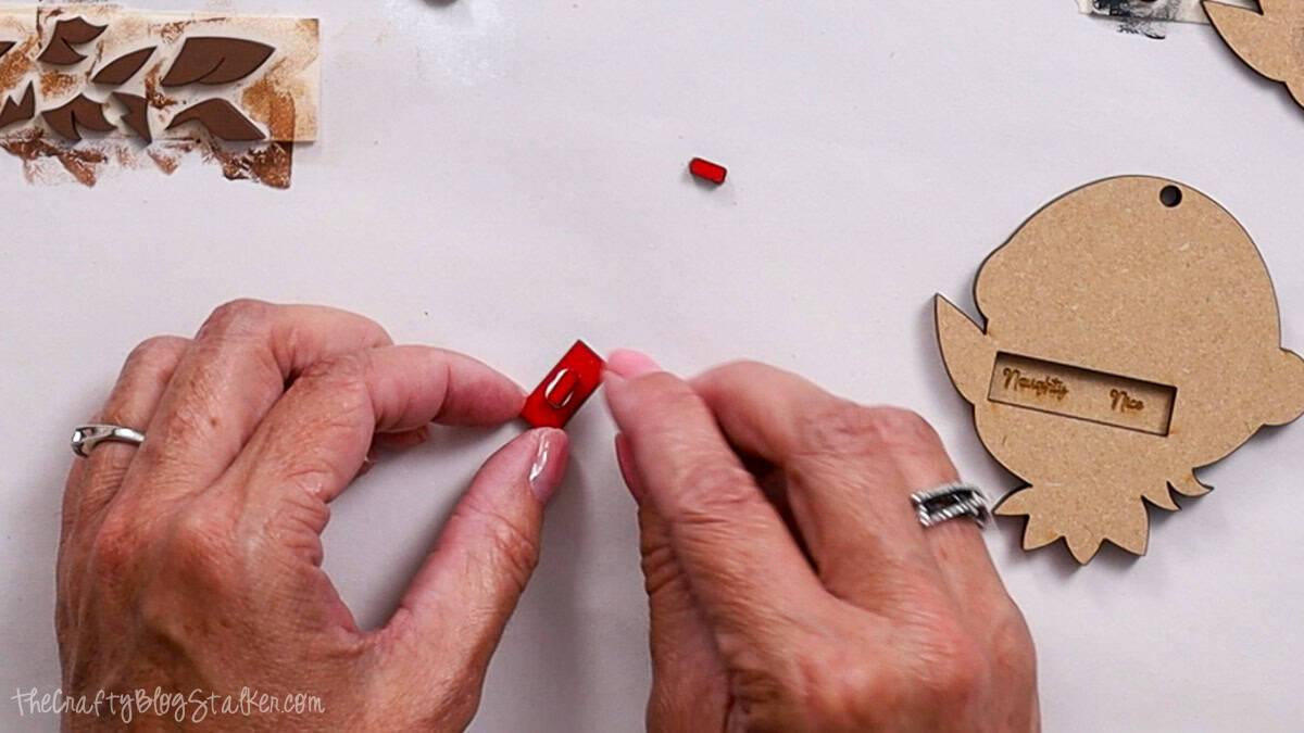 Hands assembling a small red slider piece for a laser cut elf ornament, with the ornament back showing "Naughty" and "Nice" engraved slots.