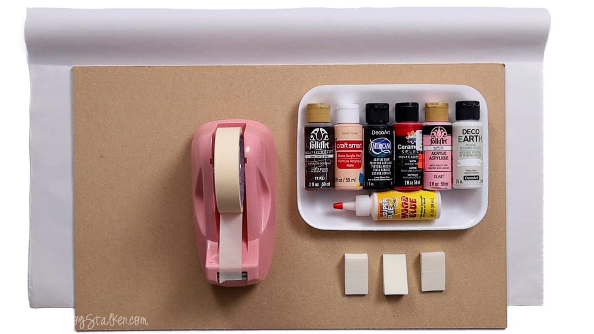 Craft supplies for a DIY project including acrylic paints, glue, makeup sponges, pink tape dispenser, and a wooden board background.