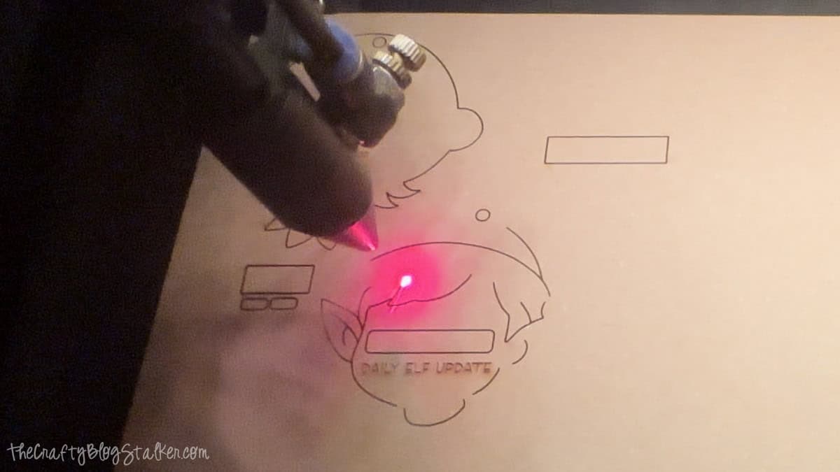 Laser cutting machine engraving elf ornament shapes on wood with a red laser beam, showing outlines and details for a DIY holiday craft project.