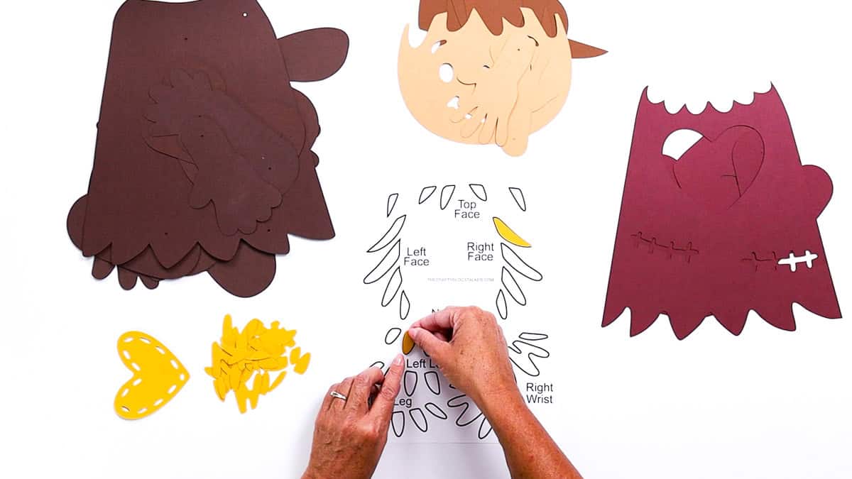 Hands arranging cut paper pieces for a DIY scarecrow craft, with a printable PDF guide showing where each part like face, hair, and clothing should be placed.