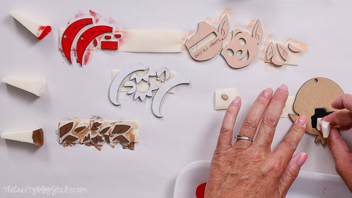 Hand painting small laser cut elf ornament pieces in red, white, black, and brown, with foam wedges and wood cutouts laid out on a crafting surface.