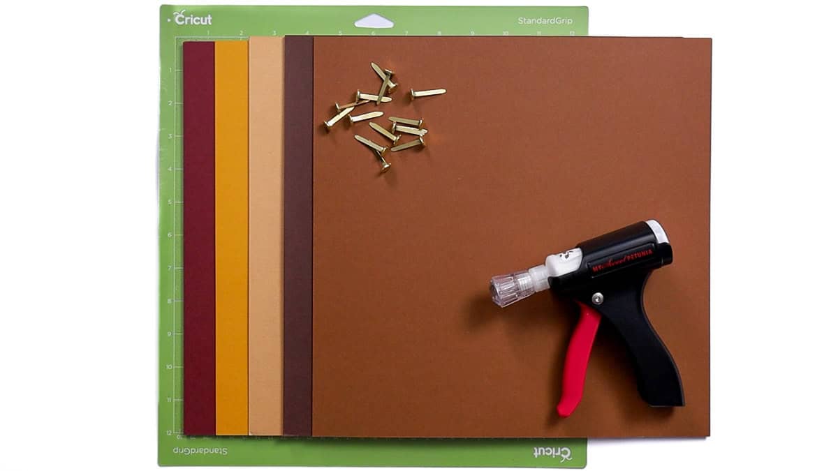 Cricut supplies for a posable scarecrow craft, including fall-colored cardstock, brads, a glue gun, and a Cricut cutting mat arranged neatly.