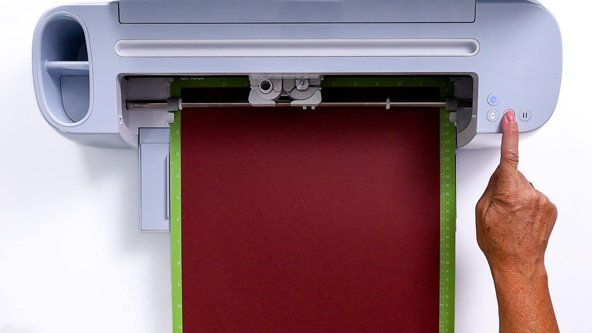A Cricut cutting machine with a green mat loaded with burgundy cardstock, ready to cut, as a hand presses the flashing start button.