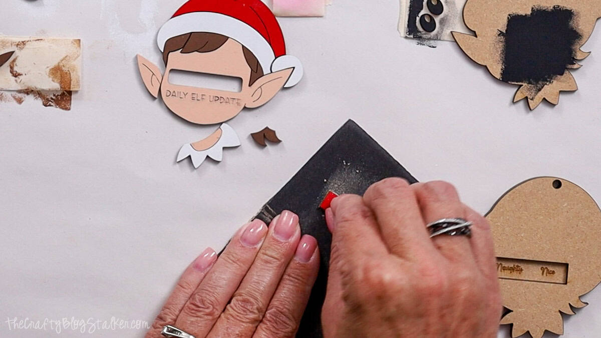Hands sanding a small red slider piece on sandpaper, with painted laser cut elf ornament parts, including hat, ears, and collar, laid out on the workspace.