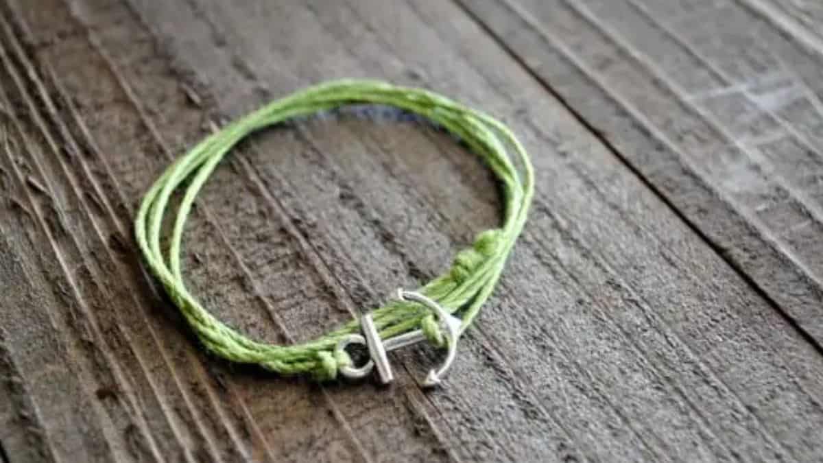 A simple handmade bracelet made of green cord with a small silver anchor charm, displayed on a rustic wooden surface.