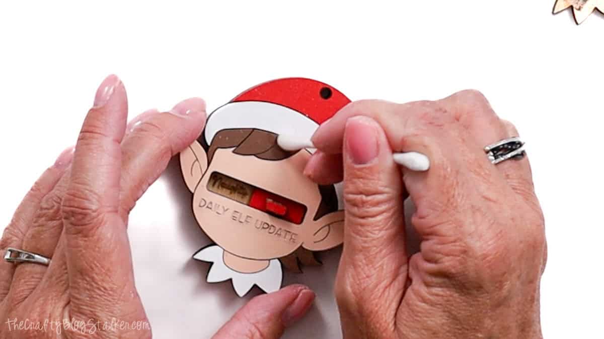 Hands assembling a painted laser cut elf ornament with a red slider showing "Naughty/Nice" and the text "Daily Elf Update" on the front.
