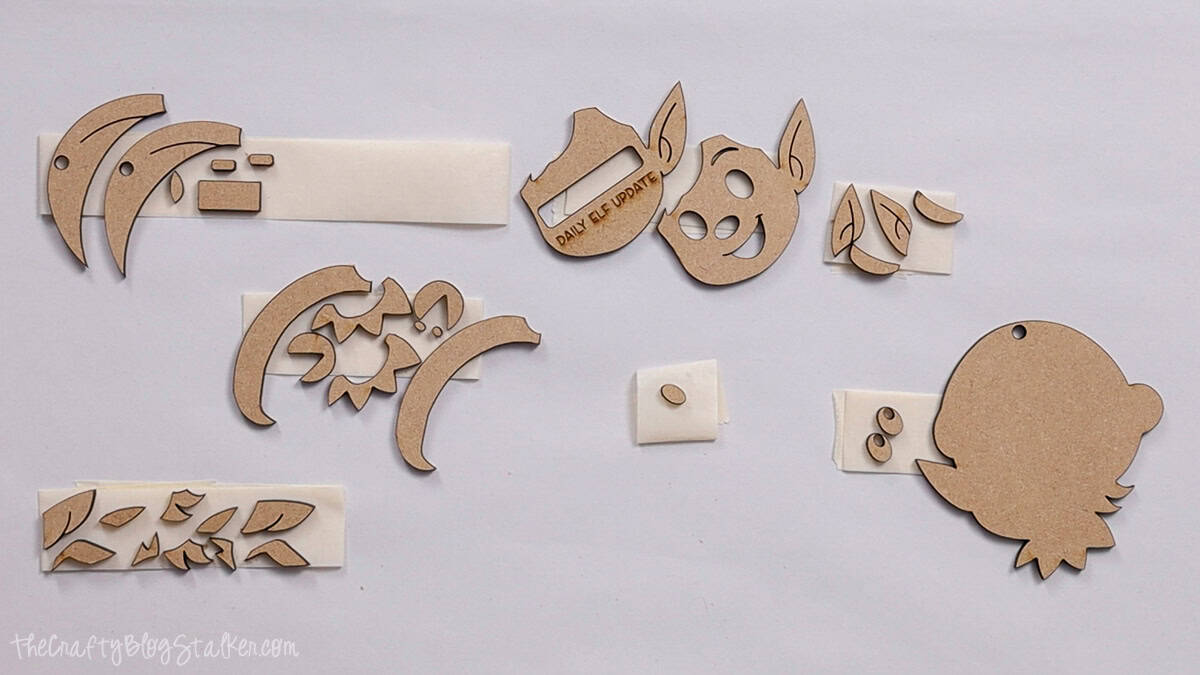 Assorted laser cut wood pieces arranged on a table, including elf face parts, ears, hat, and small shapes, ready to assemble into Christmas ornaments.