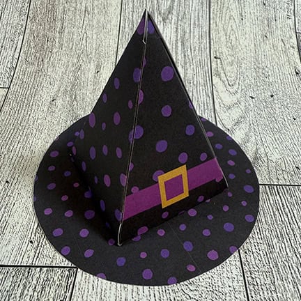 Paper craft witch hat decorated with purple polka dots and a purple belt with a yellow buckle, displayed on a wooden surface.