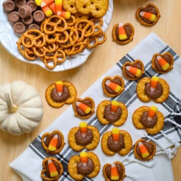 Candy corn Rolo pretzel bites arranged on a napkin and wooden table with a bowl of ingredients and white pumpkin for a festive fall treat display.