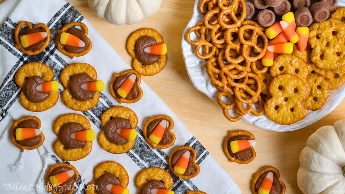 Candy corn Rolo pretzel bites arranged on a napkin and plate, surrounded by extra pretzels, Rolos, and white pumpkins on a wooden surface.