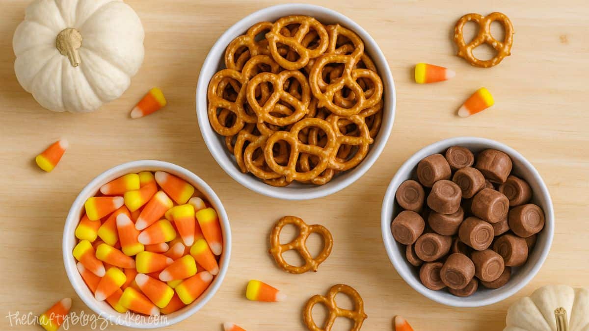 Bowls filled with candy corn, Rolos, and pretzels on a wooden surface with scattered candies and small white pumpkins for a fall-themed treat setup.