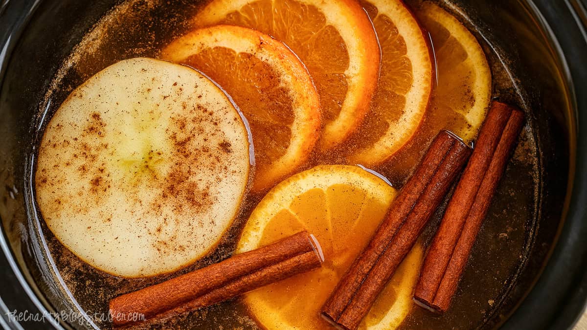 Sliced apples, oranges, and cinnamon sticks simmering in water inside a slow cooker for a fall simmer pot.