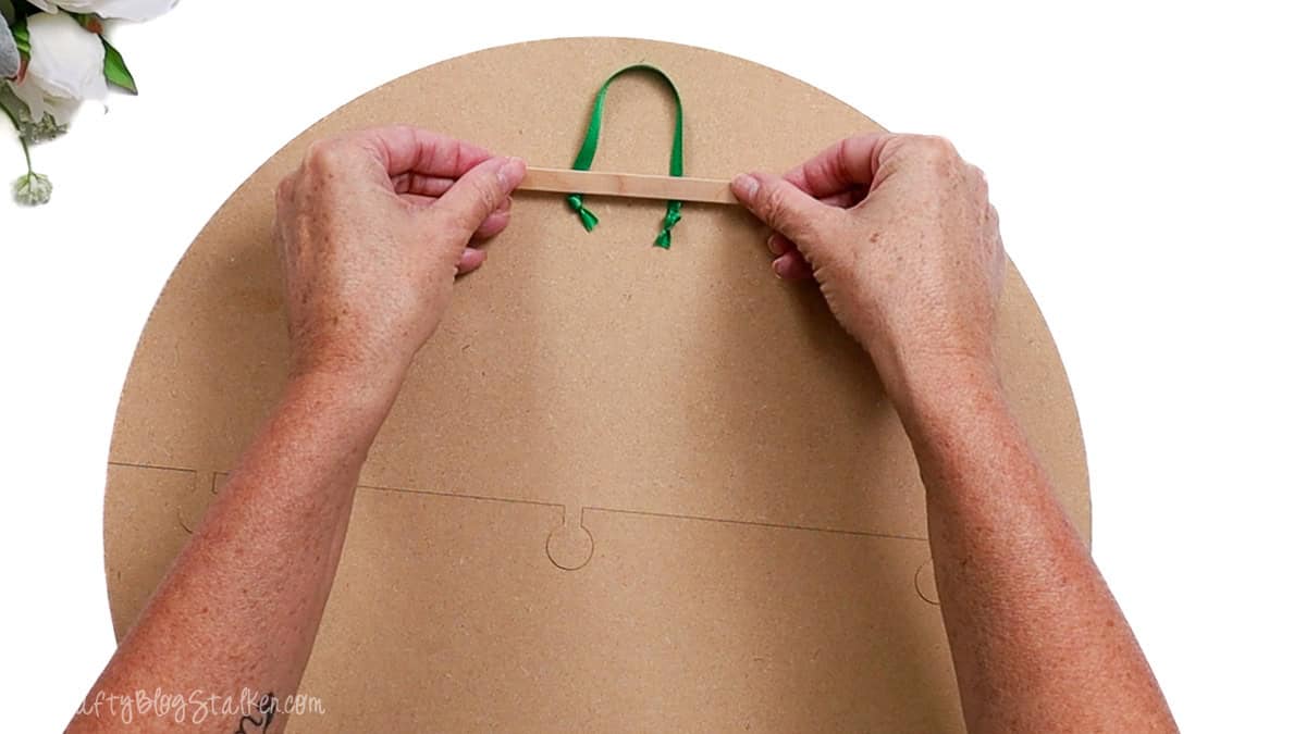 Hands gluing a green ribbon hanger to the back of a round MDF wood door hanger using a small wooden support strip.